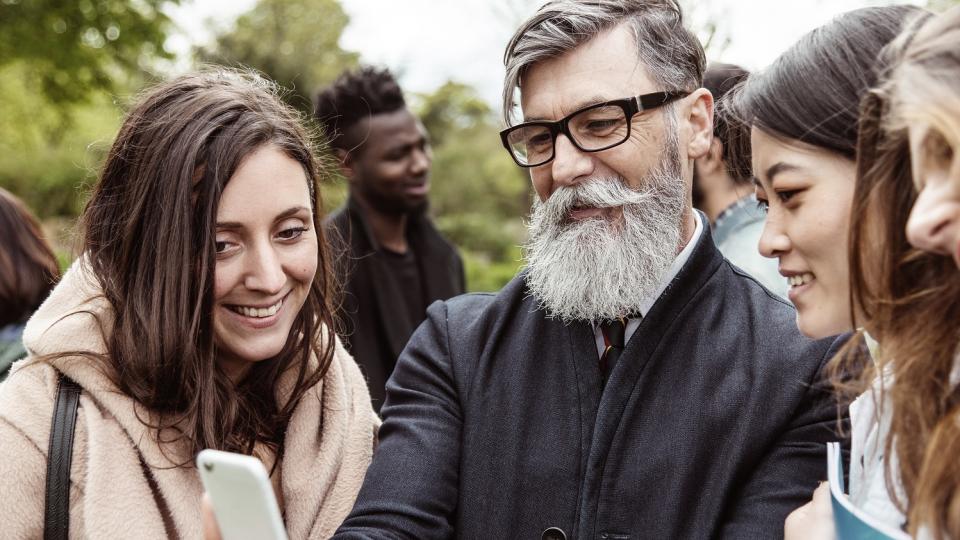 Ein älterer Mann schaut mit mehreren jungen Frauen aufs Smartphone.