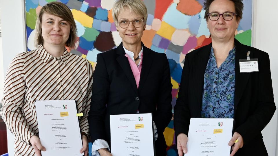 Schulministerin Dorothee Feller, Marta Grabski von Rosa Strippe e. V. und Mike Nienhaus von rubicon e. V. halten jeweils eine Vertragsurkunde in den Händen.