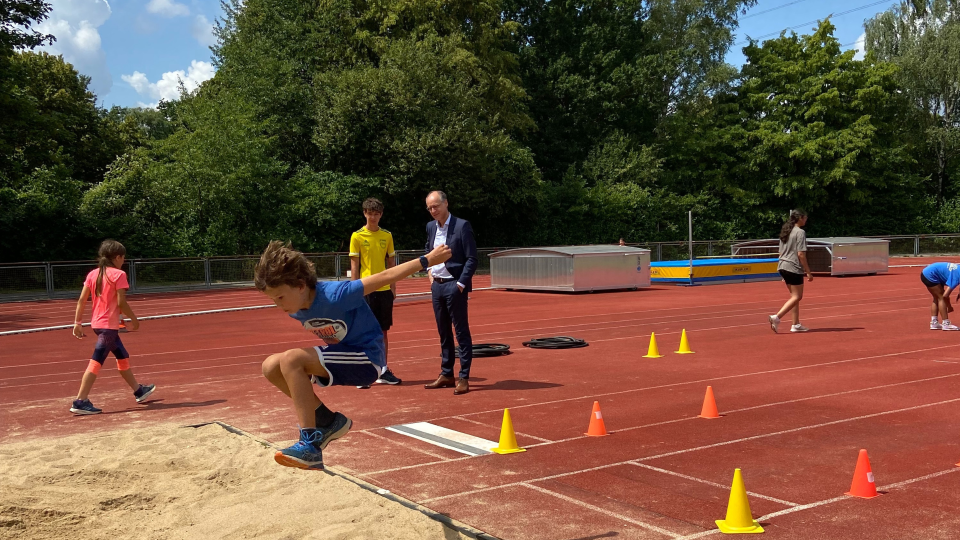 Im Vordergrund befindet sich ein Junge in Sportkleidung mitten im Weitsprung. Im Hintergrund trainieren weitere Kinder, während Staatssekretär Dr. Urban Mauer zuschaut. Im Vordergrund befindet sich ein Junge in Sportkleidung mitten im Weitsprung. Im Hintergrund trainieren weitere Kinder, während Staatssekretär Dr. Urban Mauer zuschaut.