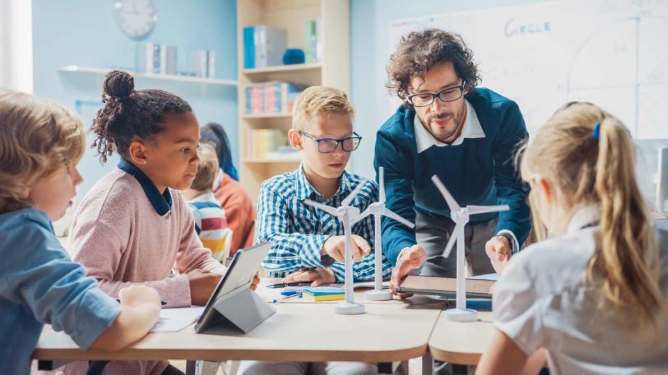 Lehrer mit Schülerinnen und Schülern in einer Unterrichtssituation. Auf dem Tisch stehen Modellwindräder, die die Kinder und der Lehrer gemeinsam betrachten.