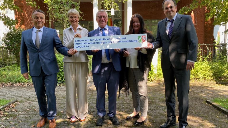 Herr Eichhorst, Ministerin Feller, Herr Schulte, Frau Guhl und Herr Geldmacher stehen mit der LAQUILA-Absenderkennung auf einem Schild in einem begrünten Innenhof Herr Eichhorst, Ministerin Feller, Herr Schulte, Frau Guhl und Herr Geldmacher stehen mit der LAQUILA-Absenderkennung auf einem Schild in einem begrünten Innenhof