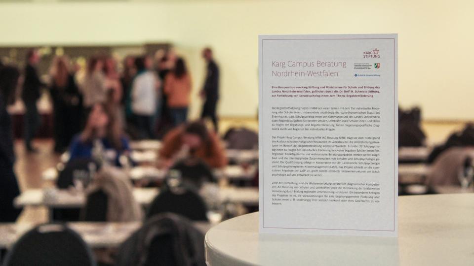 Im Hintergrund sieht man eine Tagungssituation, im Vordergrund steht eine Information zur Karg Campus Beratung auf einem Tisch. Im Hintergrund sieht man eine Tagungssituation, im Vordergrund steht eine Information zur Karg Campus Beratung auf einem Tisch.
