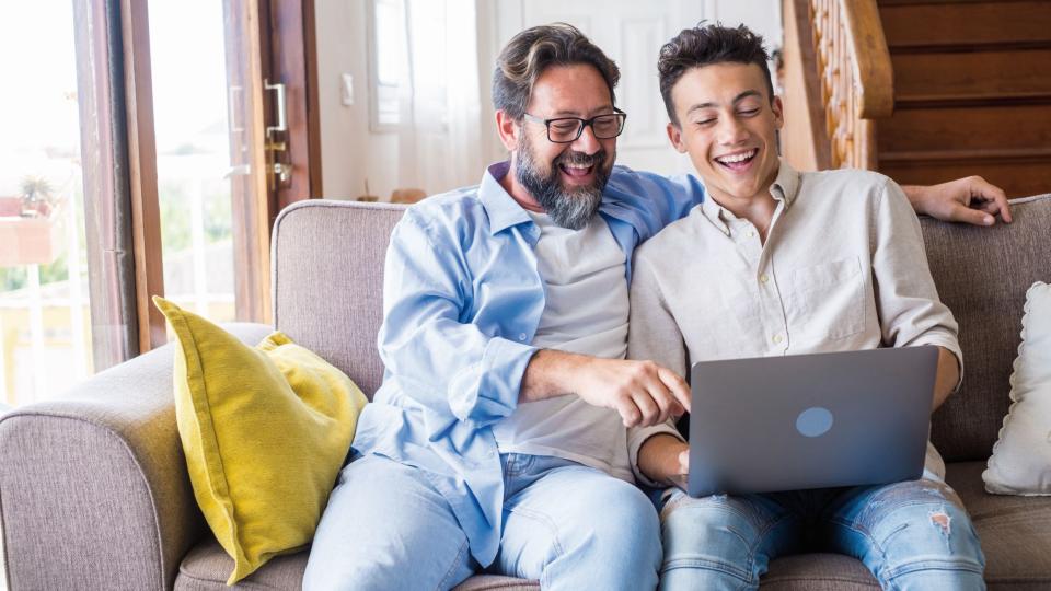 Vater und Sohn sitzen auf einem Sofa und schauen lächelnd in einen geöffneten Laptop auf dem Schoß des Sohnes.