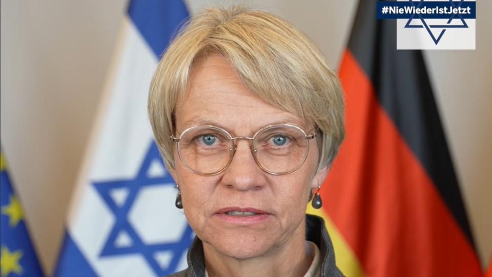 Portrait von Schul- und Bildungsministerin Dorothee Feller, im rechten oberen Bildrand ist eine Grafik des Davidsterns mit dem Schriftzug #NieWiederIstJetzt eingefügt, im Hintergrund sind die Flaggen Europas, Israels und Deutschlands erkennbar.