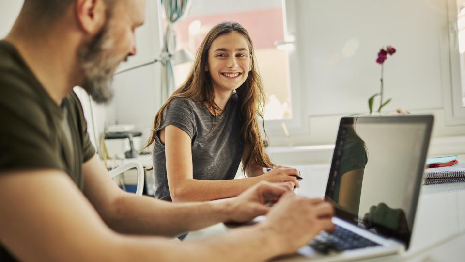 Vater und Tochter sitzen an einem Tisch, die Tochter blickt den Vater lächelnd an, während dieser etwas in einen geöffneten Laptop tippt. 