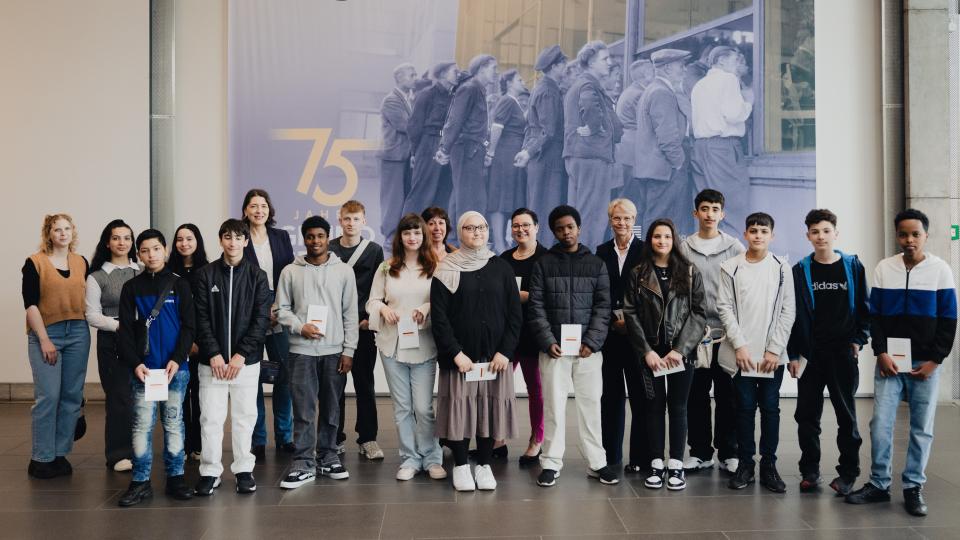 Gruppenbild: Ministerin Dorothee Feller mit Schülerinnen und Schülern der Johannes-Rau-Hauptschule Bonn im Haus der Geschichte in Bonn; im Hintergrund das Ausstellungsplakat "Ein großer Wurf - 75 Jahre Grundgesetz".