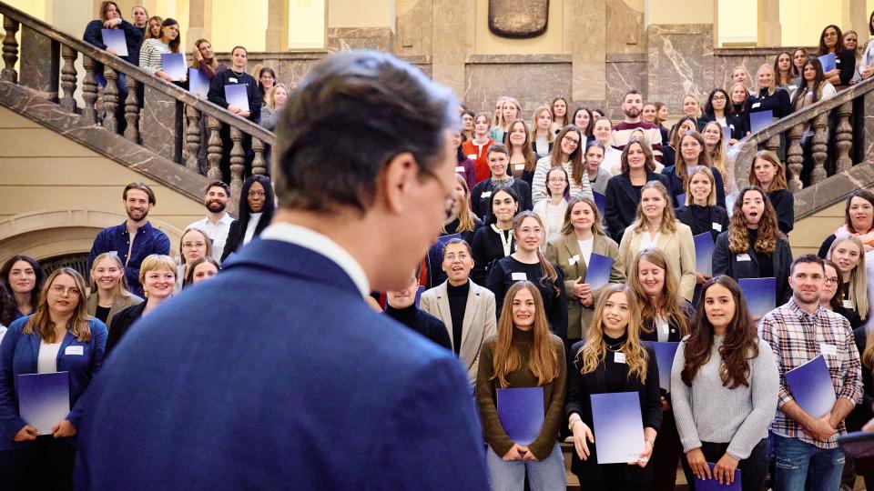 Ministerpräsident Hendrik Wüst vor einer Gruppe Lehramtsreferendarinnen und -referendare, die in einem historischen Treppenhaus mit Dokumentmappen stehen