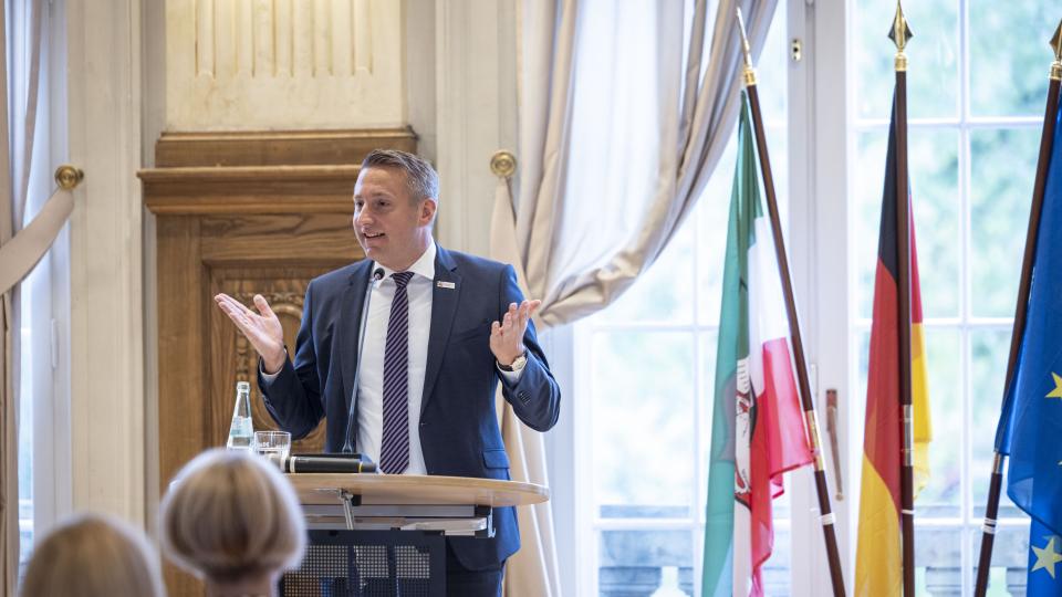 Regierungspräsident Thomas Schürmann spricht vor einer Gruppe Lehramtsreferendarinnen und -referendare, im Hintergrund die Flaggen von Europa, Deutschland und NRW