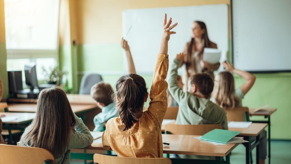 Rückansicht von Grundschülerinnen und Grundschülern, die in einem Klassenraum an ihren Tischen sitzen. Im Hintergrund ist unscharf eine Lehrerin zu erkennen. Rückansicht von Grundschülerinnen und Grundschülern, die in einem Klassenraum an ihren Tischen sitzen. Im Hintergrund ist unscharf eine Lehrerin zu erkennen.