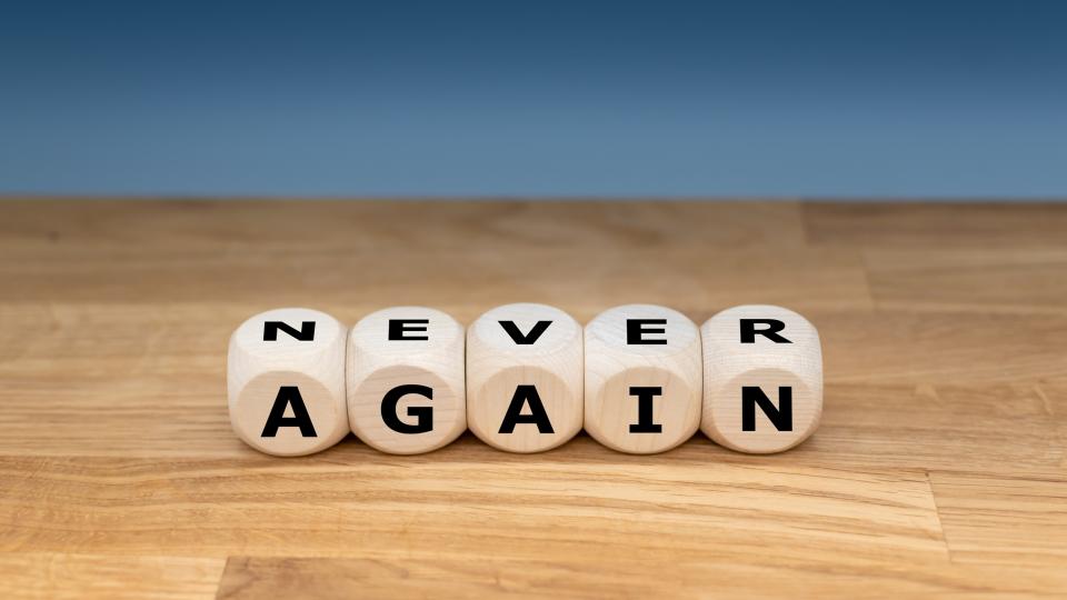 Auf einer Holzplatte liegen fünf Buchstabenwürfel, die die englischen Worte "NEVER" und "AGAIN" bilden. Auf einer Holzplatte liegen fünf Buchstabenwürfel, die die englischen Worte "NEVER" und "AGAIN" bilden.