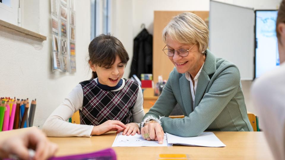 Ministerin Feller und eine Grundschülerin sitzen an einem Tisch und lesen gemeinsam in einem Buch. Ministerin Feller und eine Grundschülerin sitzen an einem Tisch und lesen gemeinsam in einem Buch.