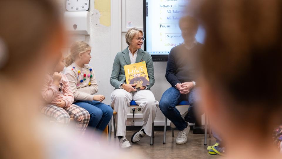 Ministerin Feller sitzt in einem Stuhlkreis mit Schülerinnen und Schülern und hält das Buch "Der Löwe in dir".