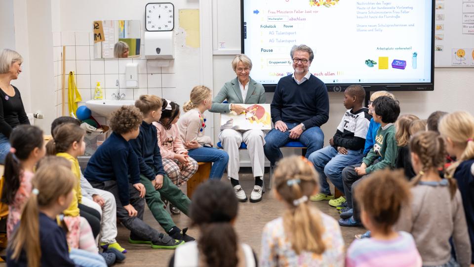 Ministerin Feller sitzt vor einer Gruppe von Schülerinnen und Schülern und liest aus dem Buch "Der Löwe in dir".
