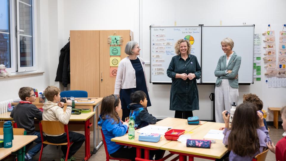 Ministerin Feller steht mit einer Lehrerin und der Schulleiterin vor einer Klasse.