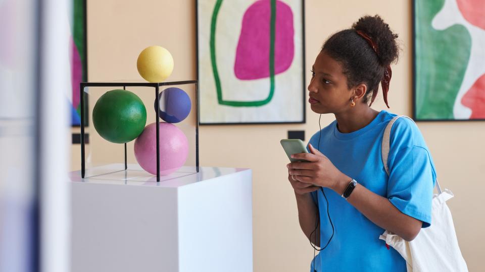 Eine Schülerin steht in einem Museum mit dem Handy in der Hand und einem Kopfhörer im Ohr vor einer Skulptur.