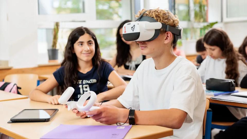 Jugendliche Schülerinnen und Schüler nutzen moderne Technologien im Unterricht. Im Fokus ein Schüler, der eine VR-Brille auf dem Kopf trägt und Controller in der Hand hält. Seine Mitschülerin schaut ihm interessiert zu.