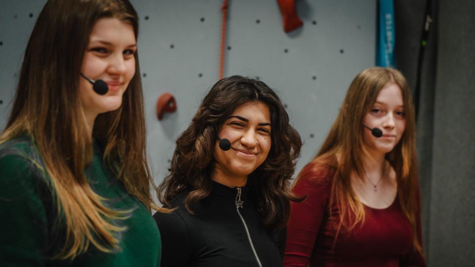 Drei langhaarige Personen  mit Headsets vor einer Kletterwand