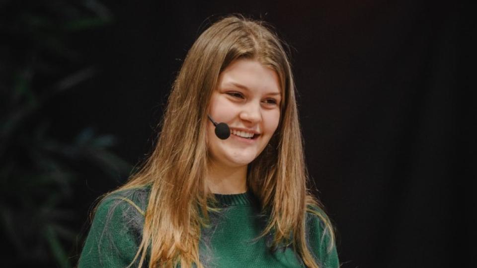 Eine langhaarige Person mit Headset steht im Publikum und hält lächelnd ein Tablet