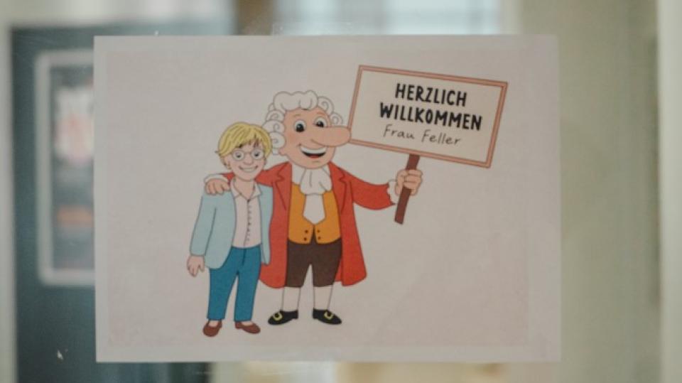 Blick in einen Schulflur mit Glastür, an der ein "Herzlich Willkommen Frau Feller"-Schild hängt, das die Ministerin und einen Schulbotschafter im historischen Gewand als Comicfiguren zeigt