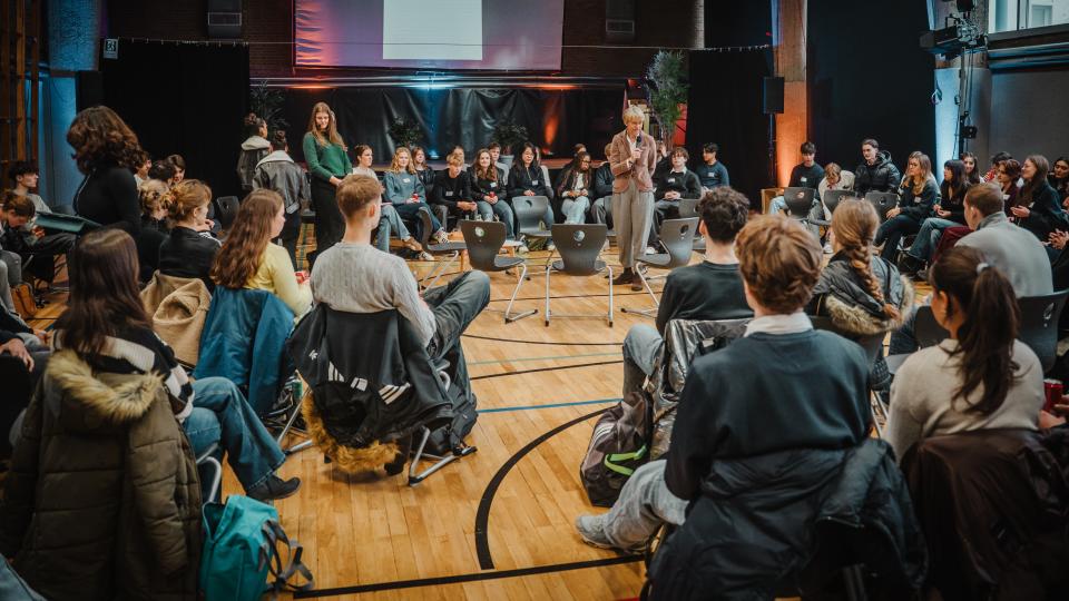 Blick in die Teilnehmendenrunde eines Fishbowl-Formats, das in einer Aula stattfindet. Im Mittelpunkt Schulministerin Feller mit einem Handmikrofon