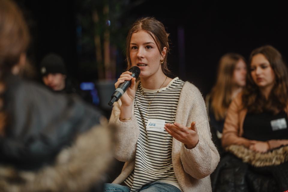 Person mit hochgesteckten Haaren spricht, umringt von sitzenden Zuhörenden, in ein Mikrofon 