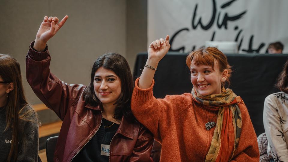 Zwei in einem Veranstaltungspublikum sitzende Jugendliche zeigen lächelnd auf