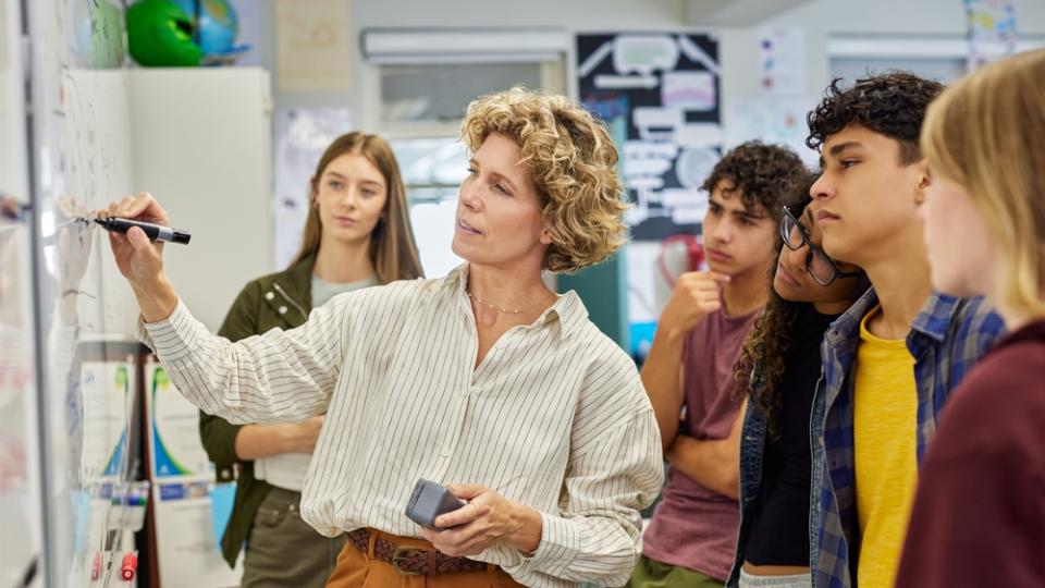 Eine Lehrerin steht an einer Tafel und erklärt den um sie herumstehenden Schülerinnen und Schülern einen Sachverhalt.