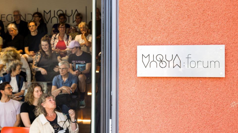 Durch die geöffnete Tür blickt man ins MiQua.forum, in dem vielen Menschen an einer Veranstaltung teilnehmen.