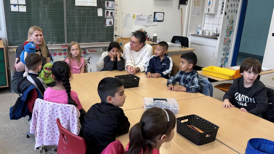 Lehrerin sitzt mit einer Handpuppe vor Kindern, die an einem Tisch sitzen.