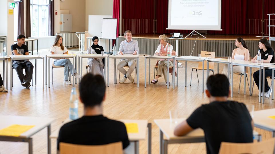 Ministerin Dorothee Feller und Jörg Schönenborn sitzen mit Schülerinnen und Schülern an im Kreis aufgestellten Tischen und sprechen miteinander.