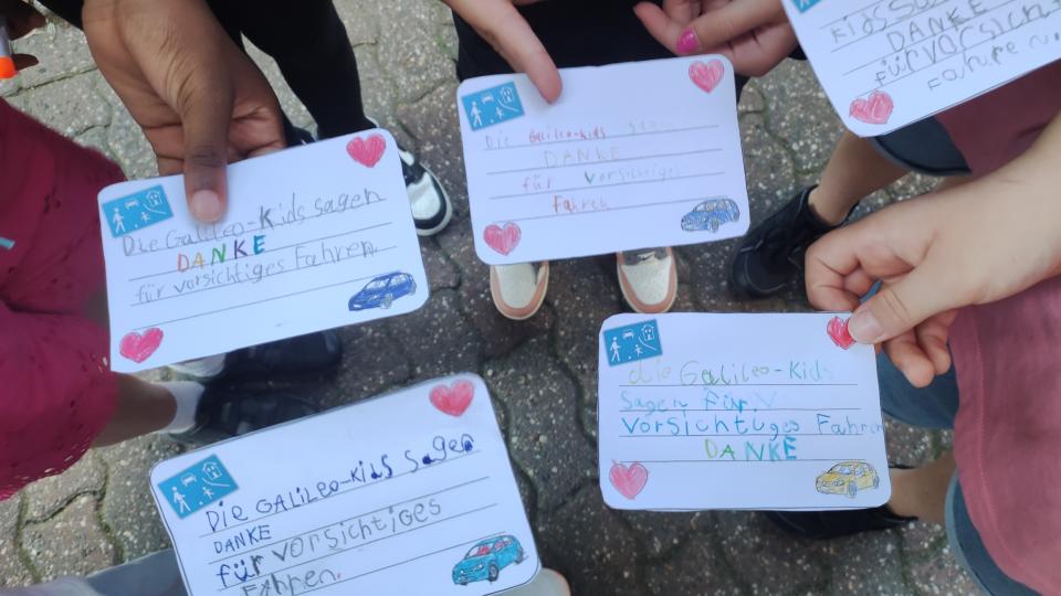 Mehrere Kinderhände halten beschriftete Karten zum Thema Straßenverkehr in die Bildmitte.