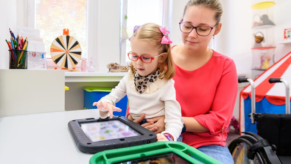 Lehrerin und Kind nutzen ein Gerät der unterstützten Kommunikation.