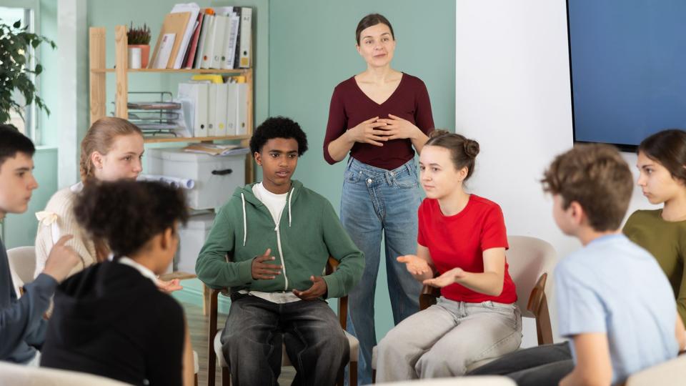Schülerinnen und Schüler sitzen im Kreis und diskutieren, eine Lehrerin beobachtet die Diskussion im Hintergrund.
