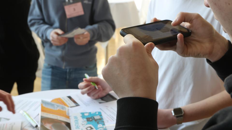 Mehrere Menschen stehen an einem Tisch, dabei macht eine Person ein Foto mit einem Smartphone.