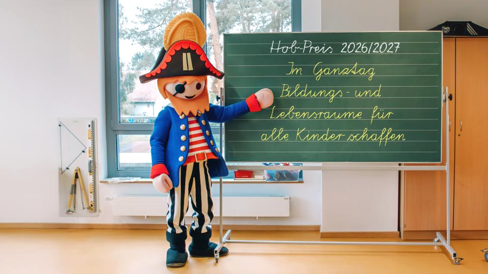 Ein Playmobil-Pirat steht an einer Tafel mit der Aufschrift "Hob-Preis 2026/27. Im Ganztag Bildungs- und Lebensräume für alle Kinder schaffen".
