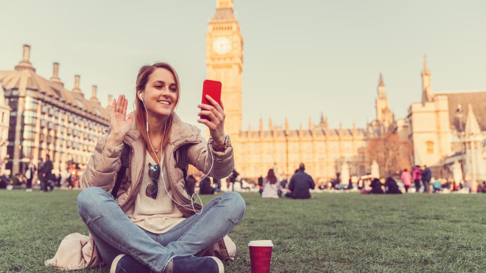 Eine Schülerin sitzt auf einer Wiese und winkt in ihr Smartphone, im Hintergrund sind die Houses of Parliament und Big Ben in London zu erkennen.