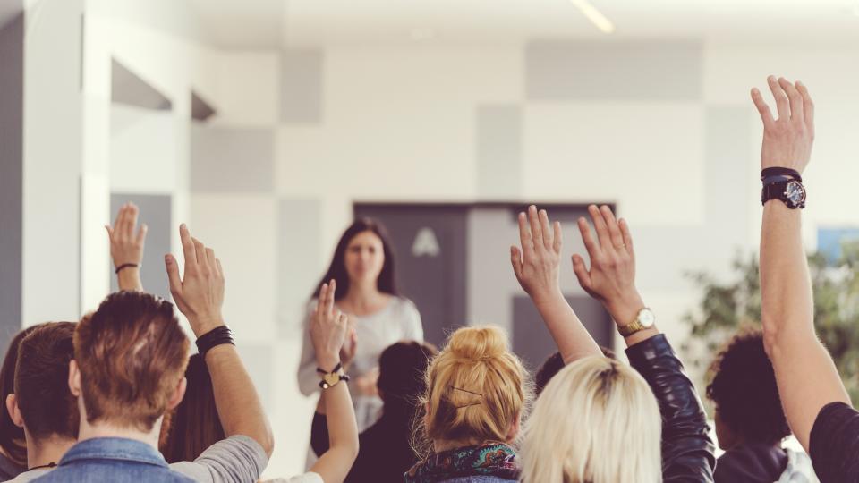 Lehrerin vor einer Gruppe Jugendlicher oder Erwachsener, viele von ihnen haben eine Hand zum Melden erhoben. Lehrerin vor einer Gruppe Jugendlicher oder Erwachsener, viele von ihnen haben eine Hand zum Melden erhoben.