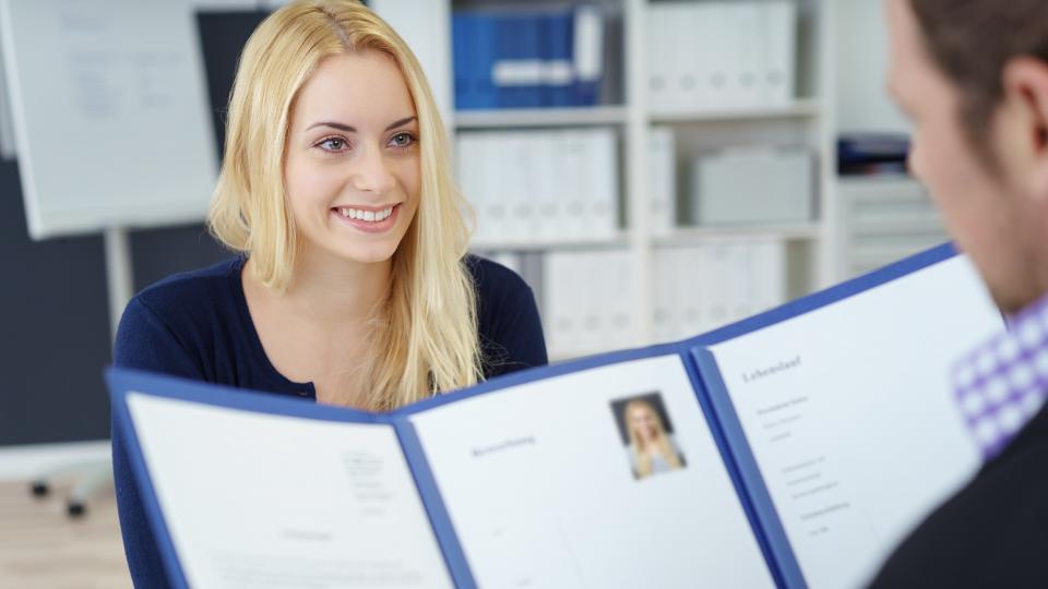 Bewerberin sitzt vor einem Mann, der ihre geöffneten Bewerbungsunterlagen anschaut. Bewerberin sitzt vor einem Mann, der ihre geöffneten Bewerbungsunterlagen anschaut.