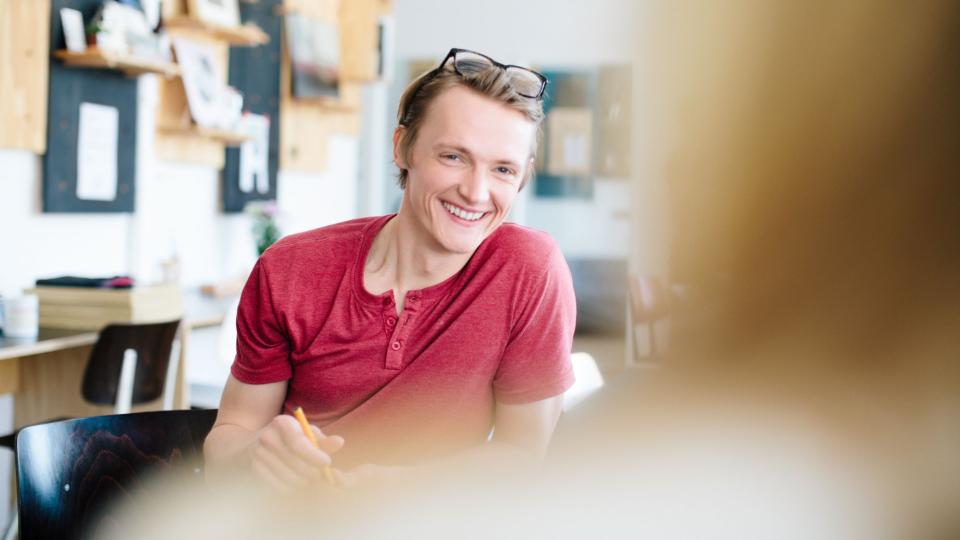 Lächelnder junger Lehrer im Gespräch mit einer Person, die man nur unscharf von hinten erkennt. 