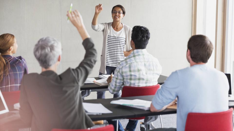 Eine Gruppe erwachsener in einer Besprechungssituation. Ein Zuhörer hat die Hand erhoben, die Moderatorin erteilt ihm das Wort.