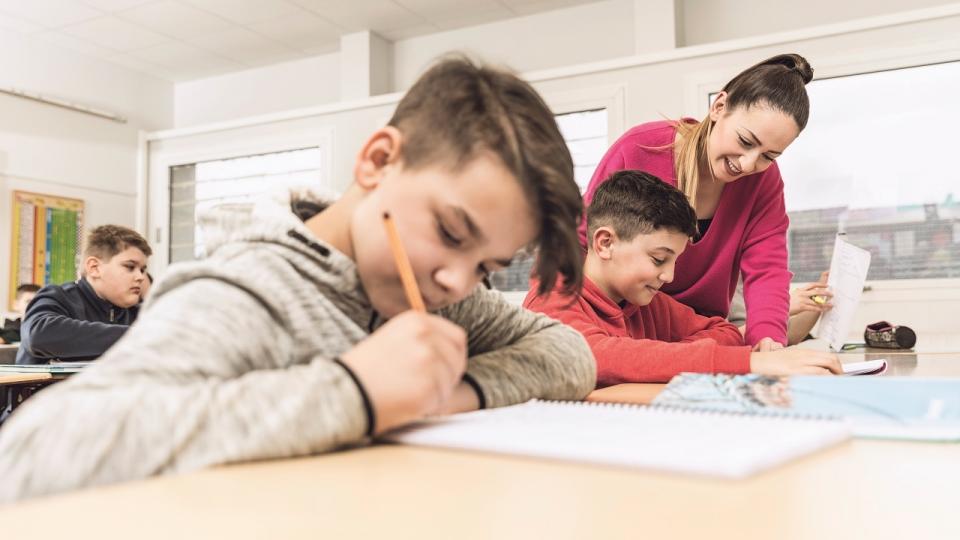 Lehrerin erklärt einem Schüler Unterrichtsstoff in einer Unterrichtssituation. Im Vordergrund sitzt ein anderer, schreibender Schüler.