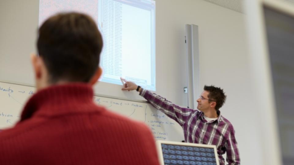 Unterrichtsituation mit digitalen Medien: Lehrer erklärt etwas an einem Smartboard, im Vordergrund steht ein Computerbildschirm.