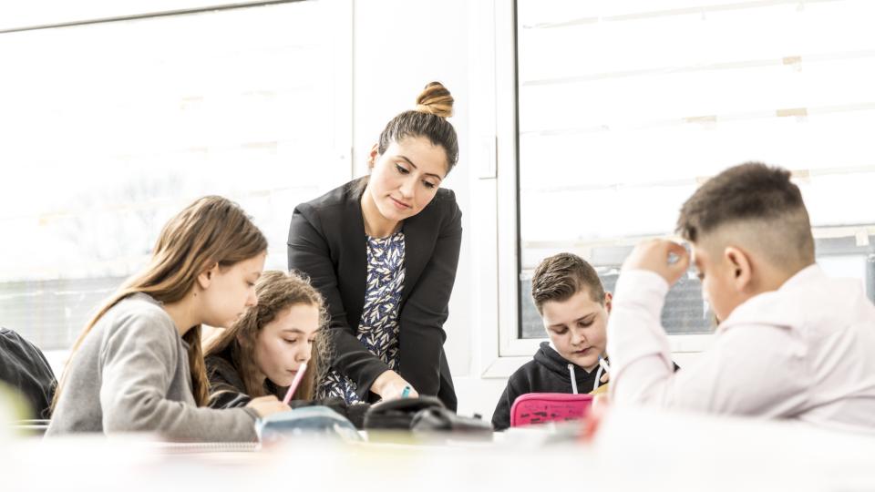 Lehrerin beugt sich erklärend über einen Tisch, an dem Schülerinnen und Schüler lernen. Lehrerin beugt sich erklärend über einen Tisch, an dem Schülerinnen und Schüler lernen.
