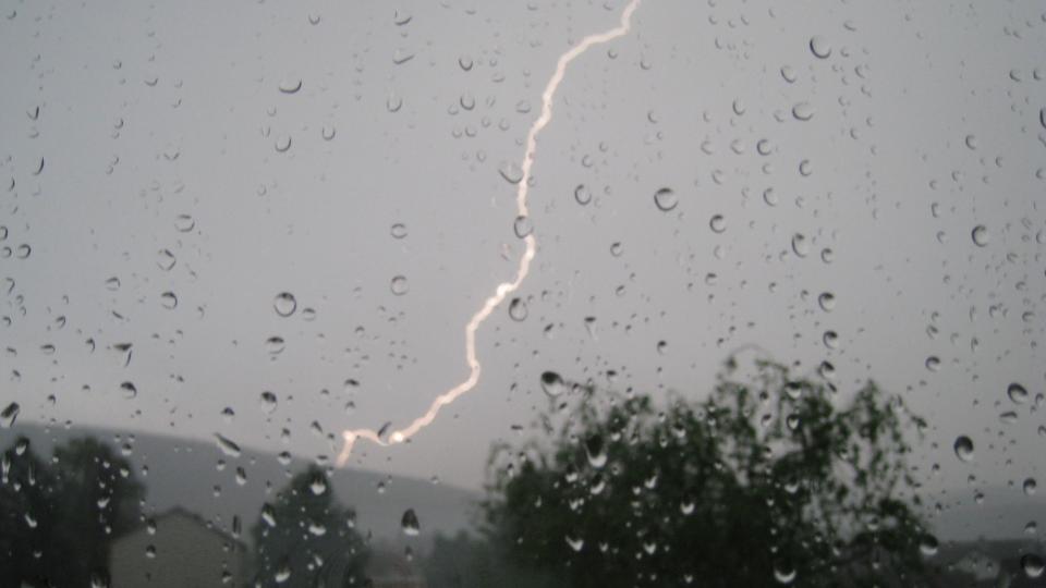 Sicht durch eine Fensterscheibe auf ein Unwetter mit Regen und Blitz, Bäume und ein Haus sind auszumachen.