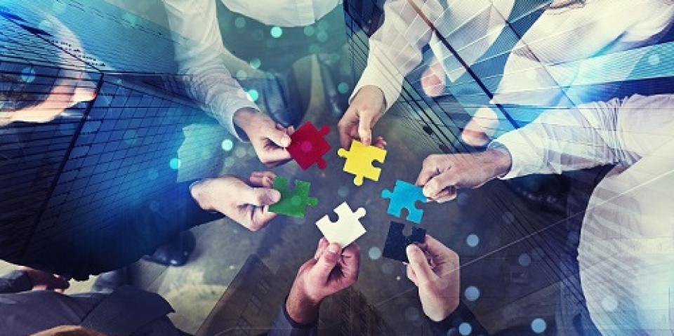 Sechs Hände halten jeweils ein Puzzleteil in die Mitte. Leicht verfremdet ist über dieses Bild eine Vogelperspektive sich spiegelnder Hochhäuser gelegt.