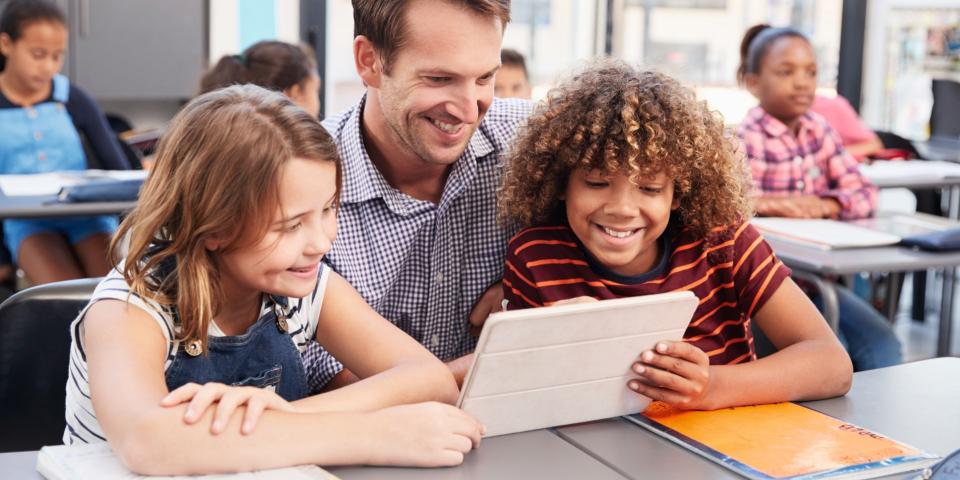Lehrer, eine Schülerin und ein Schüler blicken in einem Klassenraum gemeinsam auf ein Tablet (Computer). 