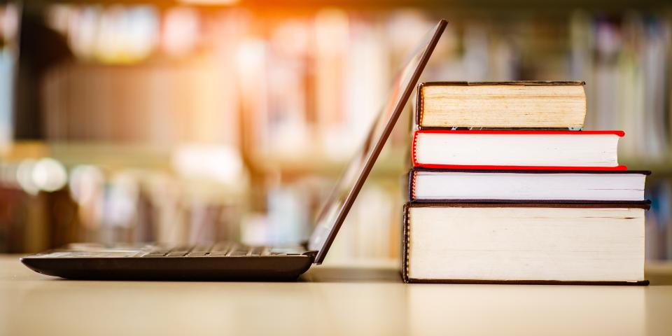 Geöffneter Laptop neben einem Bücherstapel, im Hintergrund ein Bücherregal.