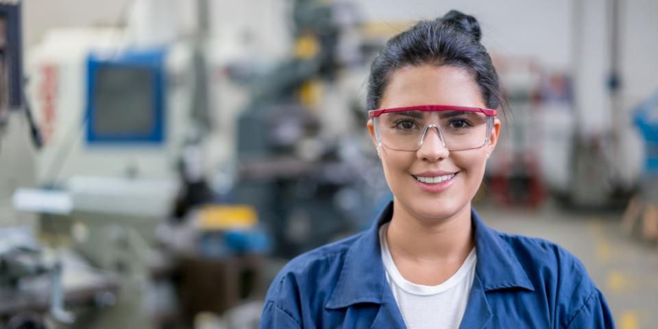 Junge Frau in Arbeitskleidung und Schutzbrille, im Hintergrund Maschinen.