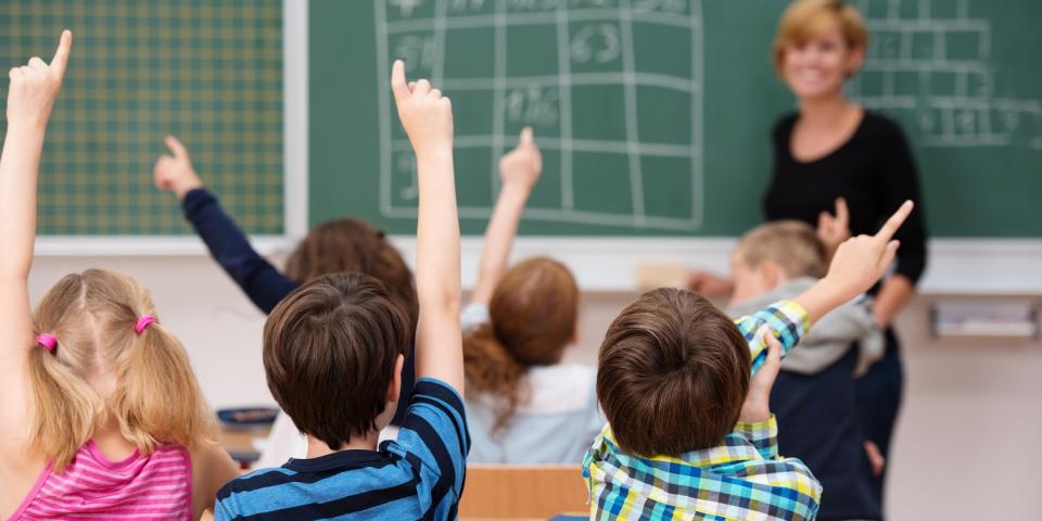Grundschülerinnen und -schüler melden sich, im Hintergrund eine Tafel mit Rechenaufgaben