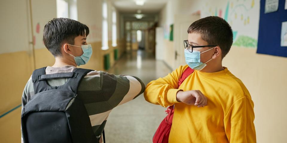Zwei Alltagsmasken tragende Schüler begrüßen sich mit einem Ellenbogencheck auf einem Schulflur.
